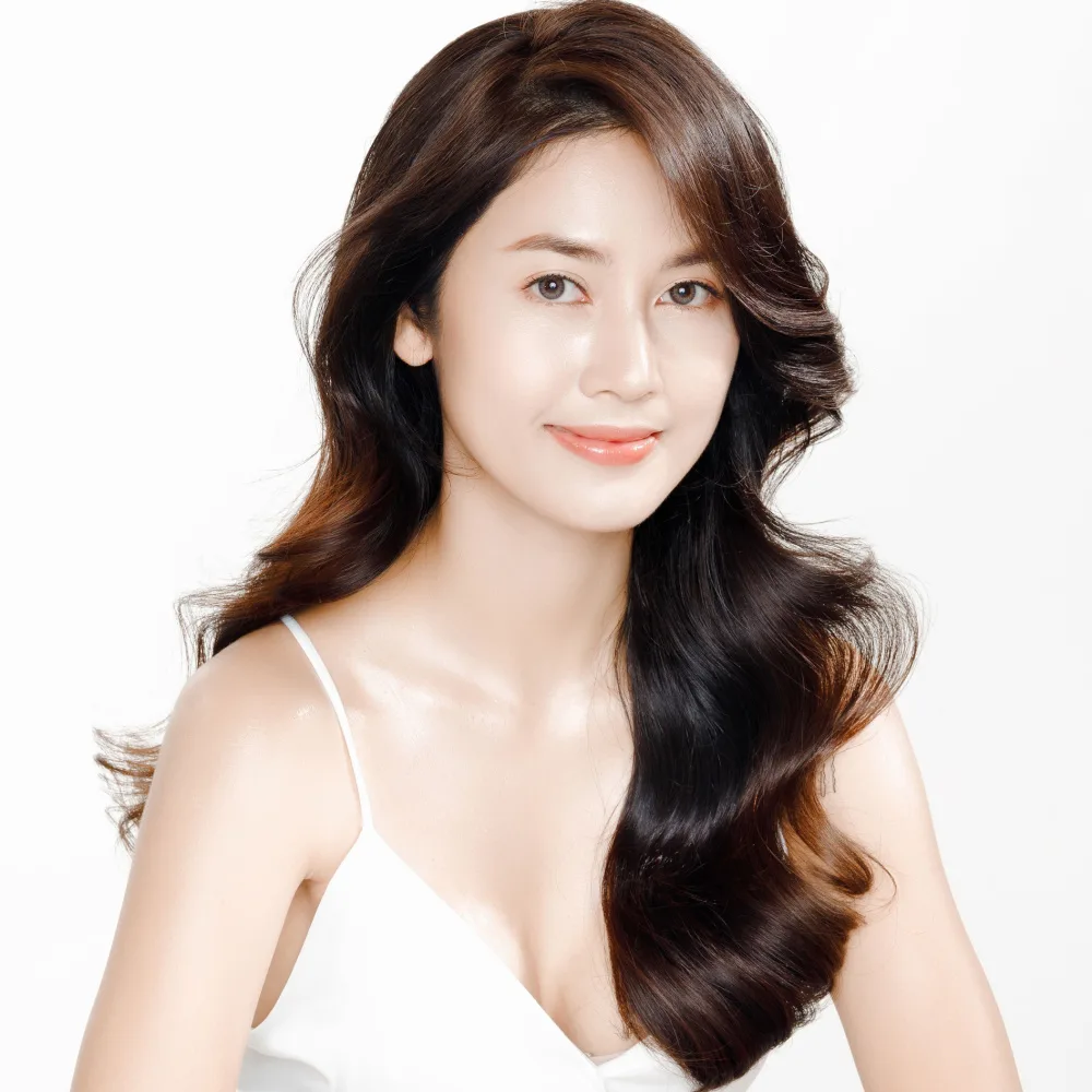 Woman smiles wearing white tank top with Asian long hair styled in big waves with side part in front of white background