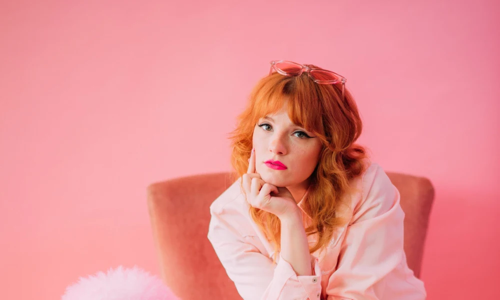 Redhaired woman sits in chair in pink room with her chin resting on her hand wearing a long shag haircut with sunglasses on her head