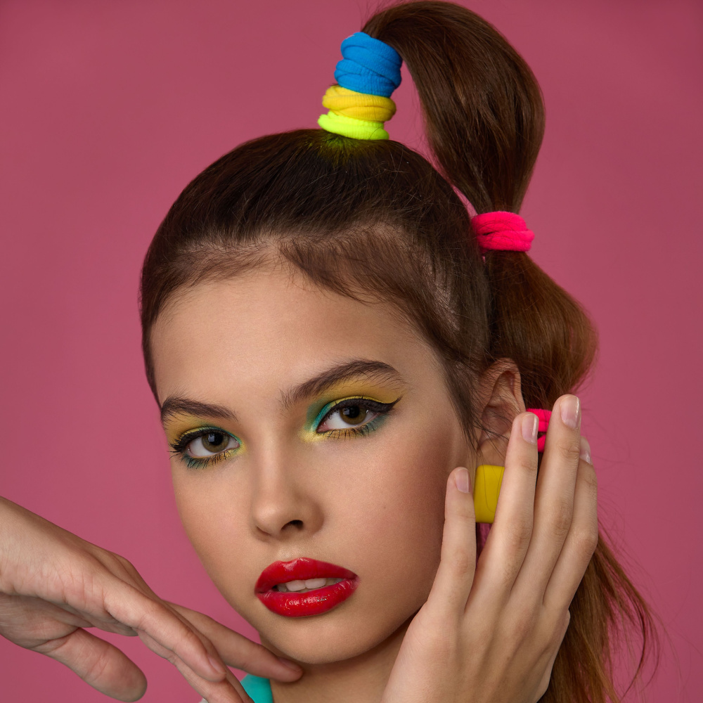 Close up view of brunette woman with rainbow colored elastics wearing 1980s bubble braid hairstyle in front of pink wall with hand on her face