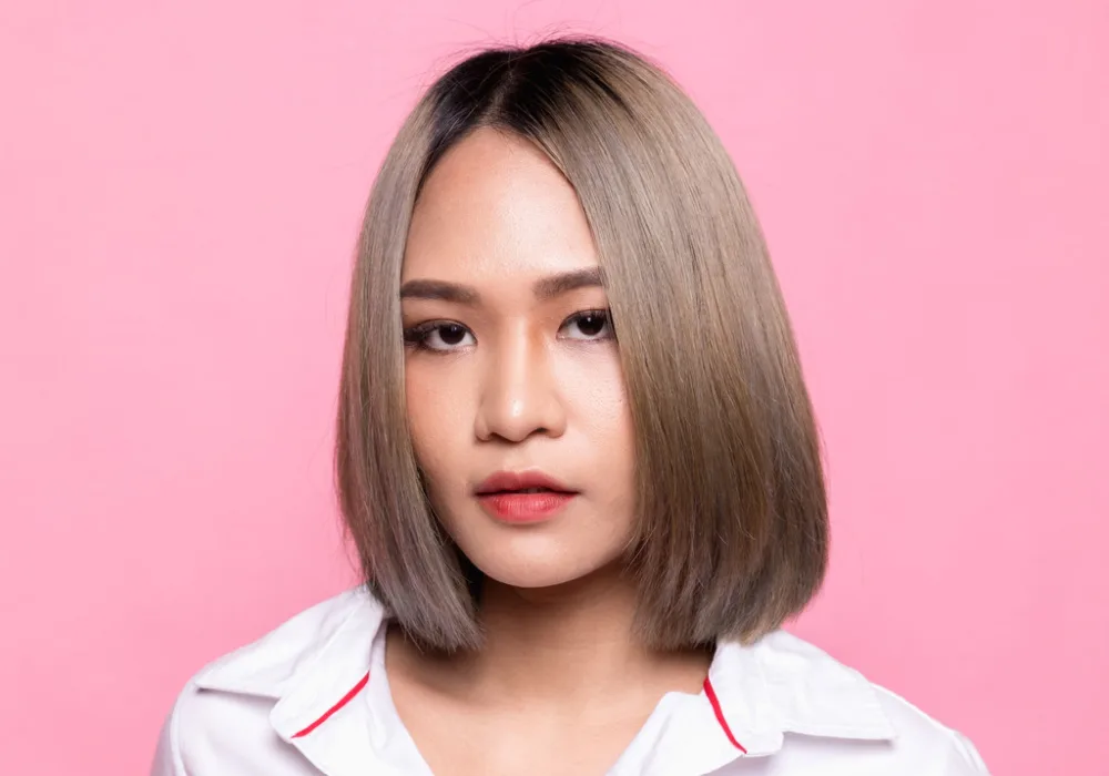 Asian woman with long bob hair shows a dark ash shade of blonde color in front of a pink background