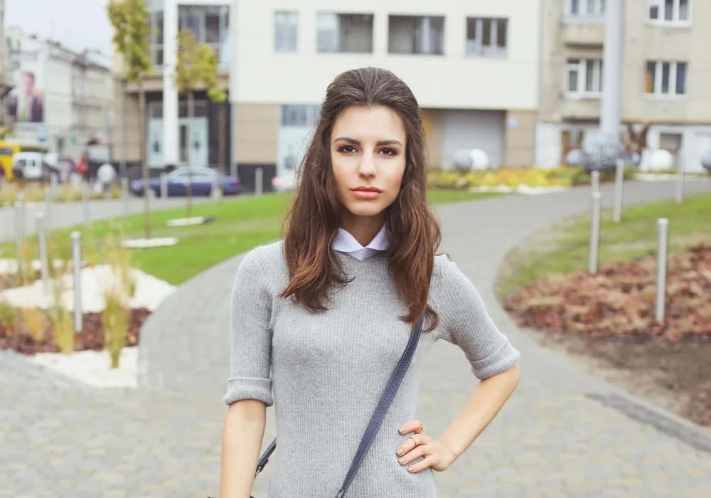 Brunette woman in gray knit dress with crossbody purse models one of her half up half down hairstyles outdoors