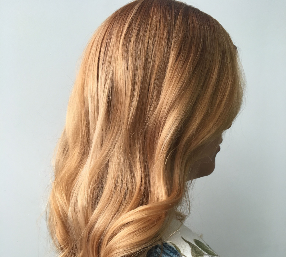 Woman faces away from the camera from a back side view with one of the red-toned shades of blonde in a strawberry blonde color