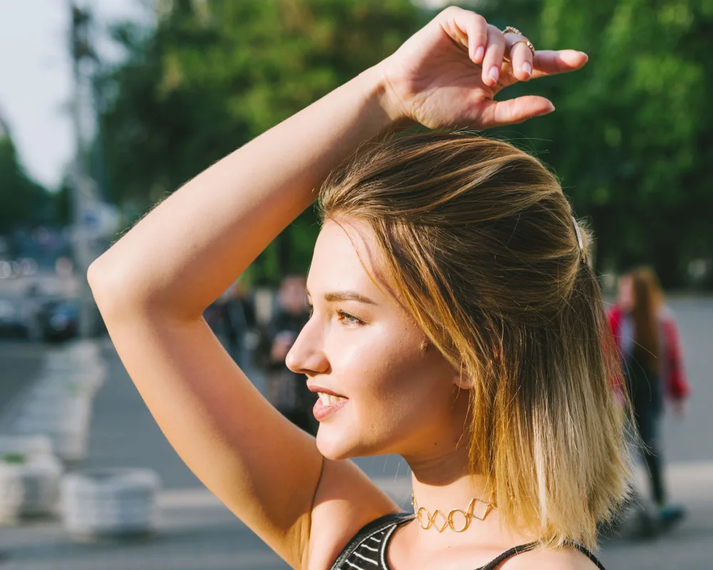 Woman with blonde balayage hair wears one of the easiest half up half down hairstyles with face-framing strands and arm raised over her head