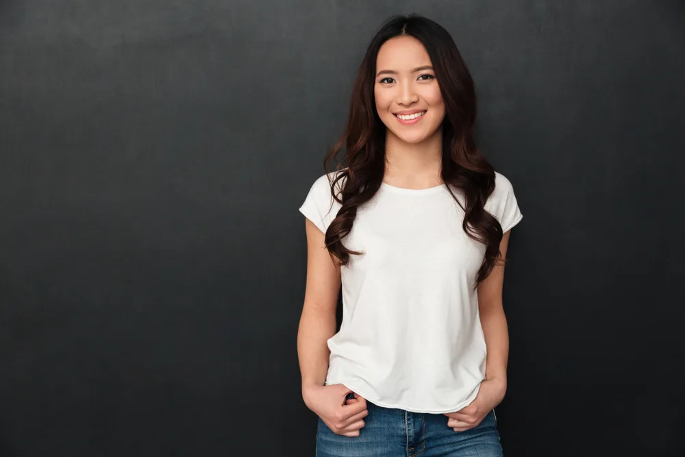 Happy Asian woman with mid back length hair wears a white shirt and jeans in front of a dark gray background with hands through her belt loops