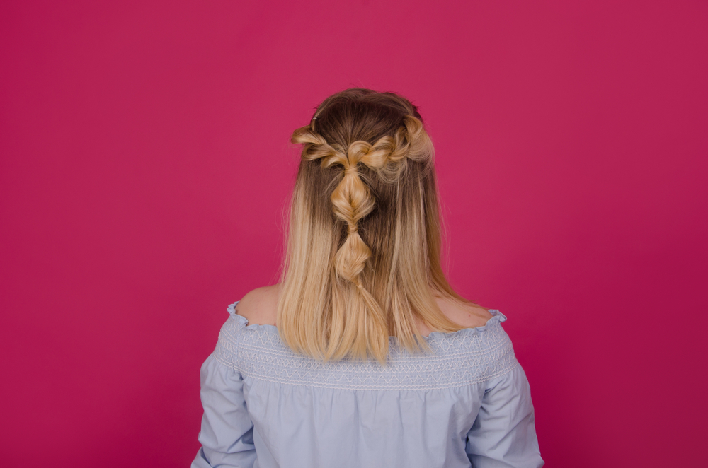 Rear view of woman with blonde hair styled in inverted bubble braids and half up twists standing in front of pink wall