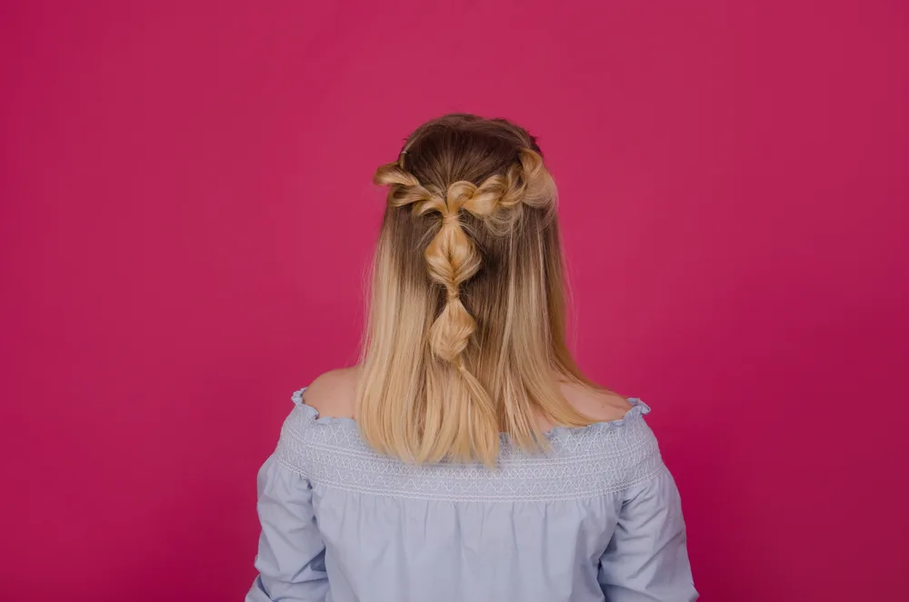 Rear view of woman with blonde hair styled in inverted bubble braids and half up twists standing in front of pink wall