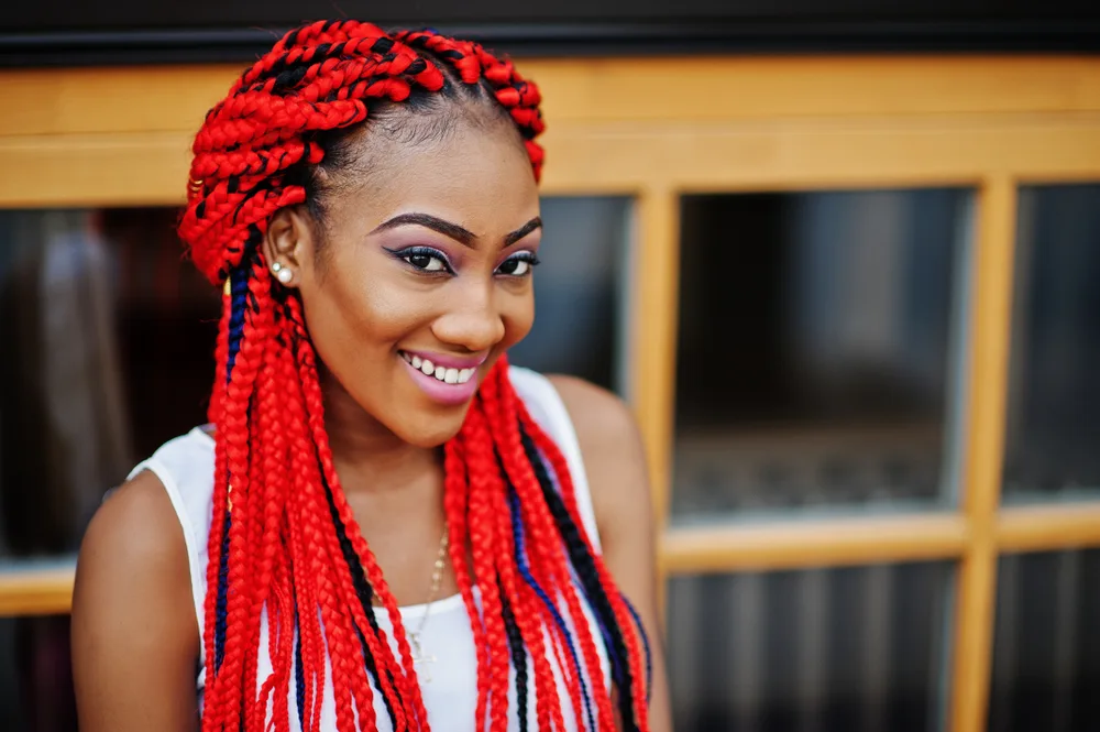 African American woman smiles in front of windows wearing bright red jumbo box braids half up half down hairstyle