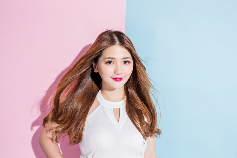 Asian woman dressed in cutout white dress in front of split pink and blue background wears brown hair color with long hair
