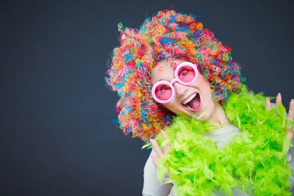 Woman wears a colorful afro curly wig with green feather boa in front of dark background while smiling in big pink sunglasses