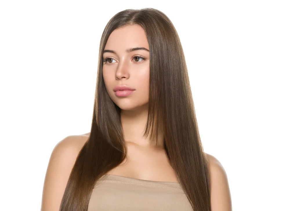 Woman with brown hair wears a tan tank top in front of white background with long straight hair to show one of the best heart shaped face haircuts