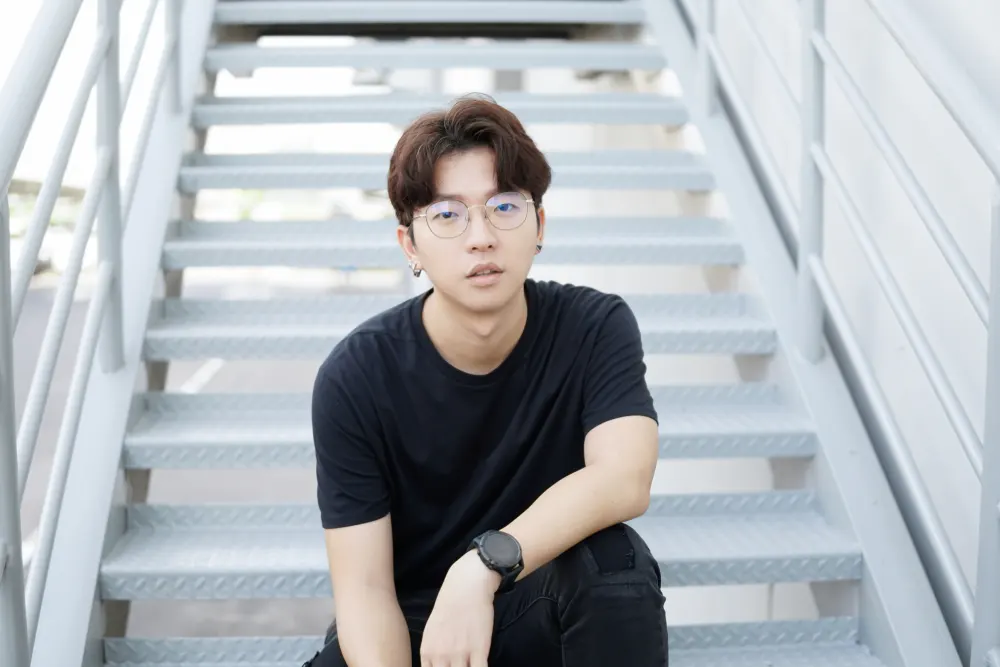 Man wearing glasses and black clothing sits on steps to show off one of many Korean men hairstyles with a curtains style