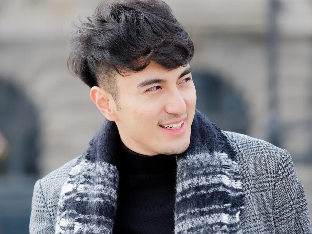 Smiling Korean man wears a coat and scarf in the city with one of the best Korean men hairstyles, a voluminous curly look with burst fade