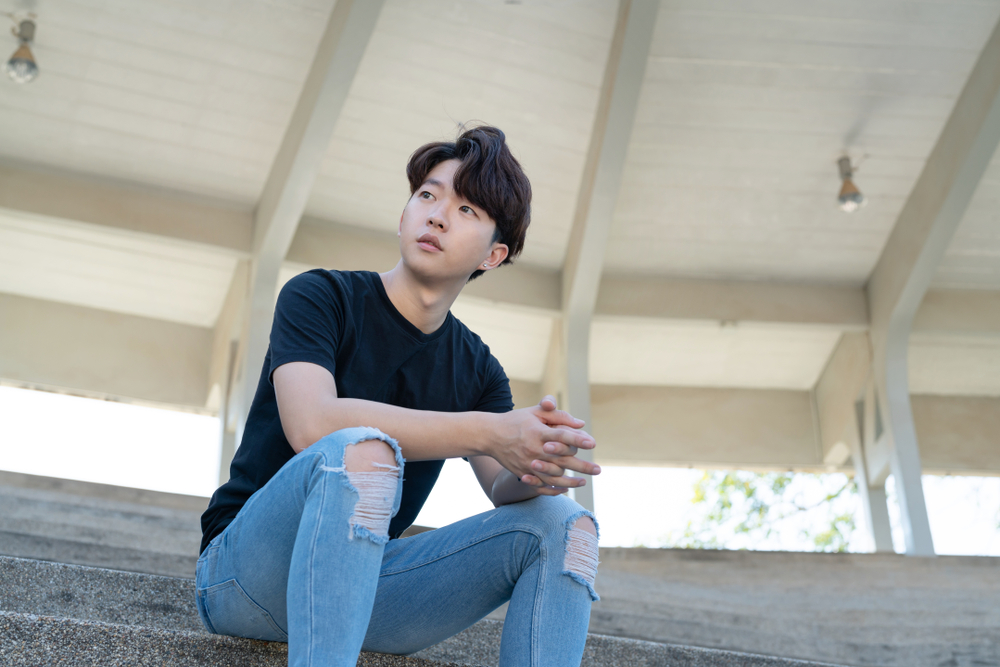 Asian man sits on steps in bandstand with casual t-shirt and jeans while wearing one of the top Korean men hairstyles with lots of volume