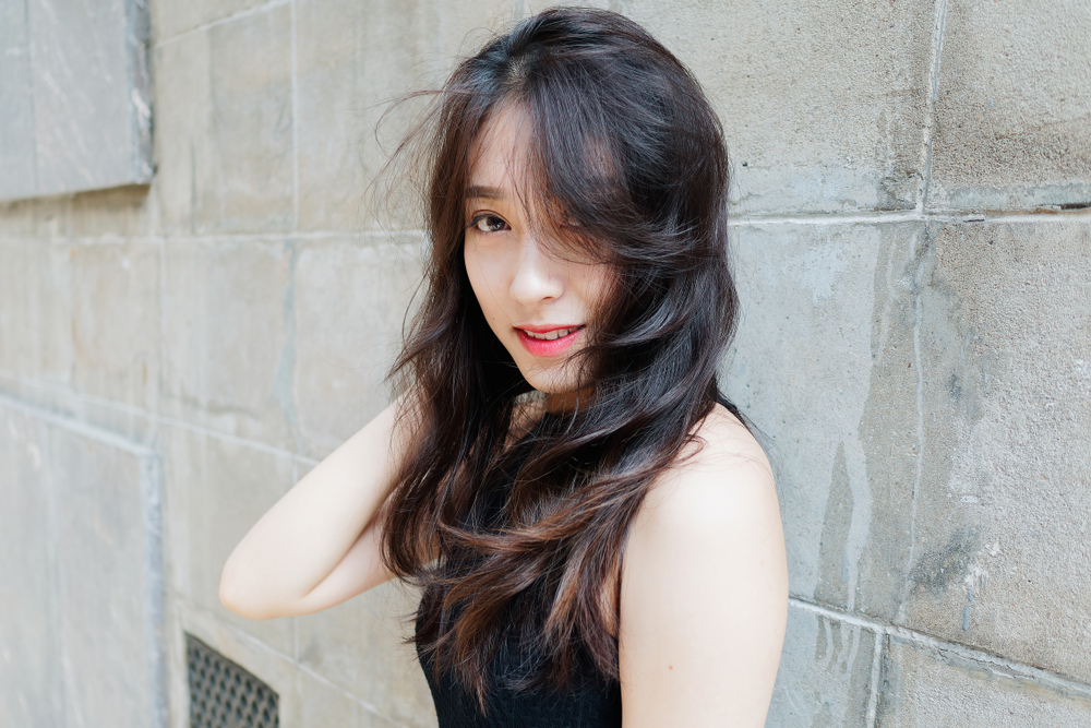 Young Asian woman in sleeveless black top stands in front of stone wall after running her hands through her long shag haircuts' length