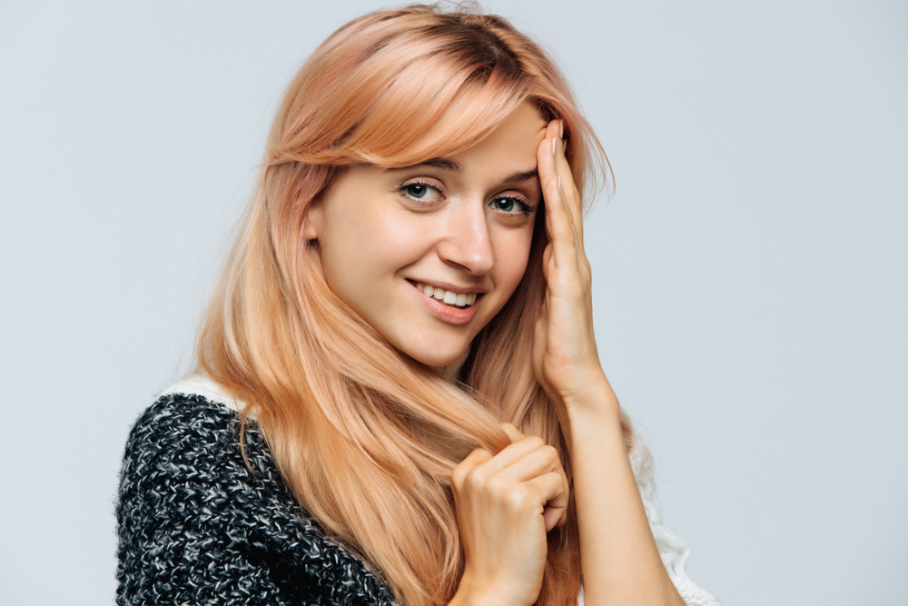 Woman smiles with hand touching her forehead to show one of the unique shades of blonde in a rose gold color with long hair and side bangs