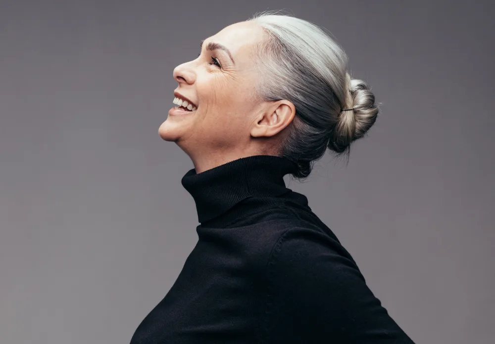 Woman holding her head up in the air and wearing a black turtleneck as part of a roundup of the best medium length layered hairstyles for women over 50