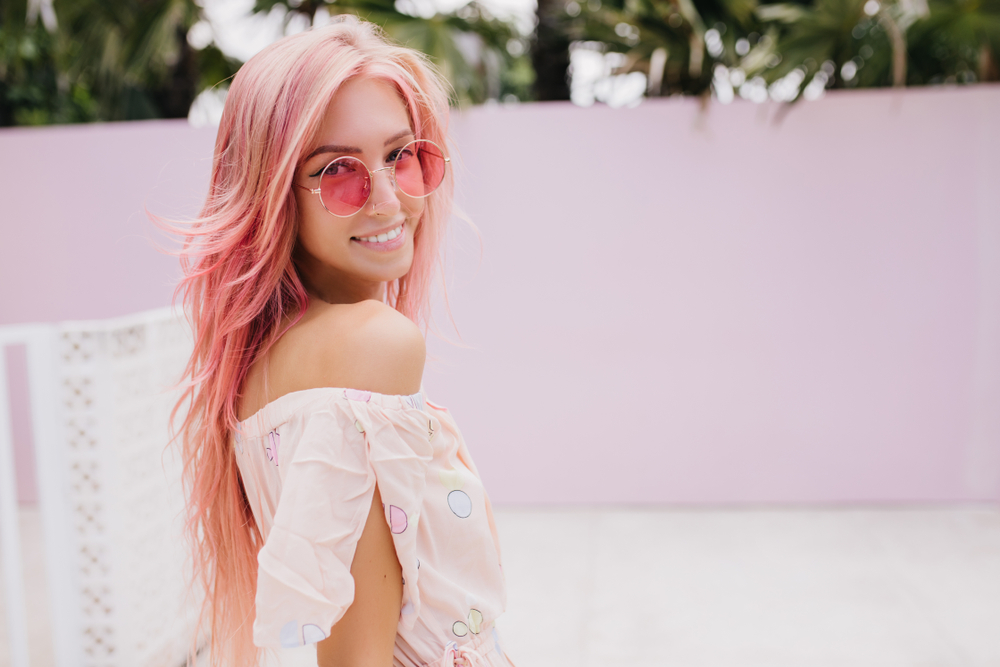 Woman with light pink hair that's almost blonde looks over her right shoulder and smiles wearing sunglasses