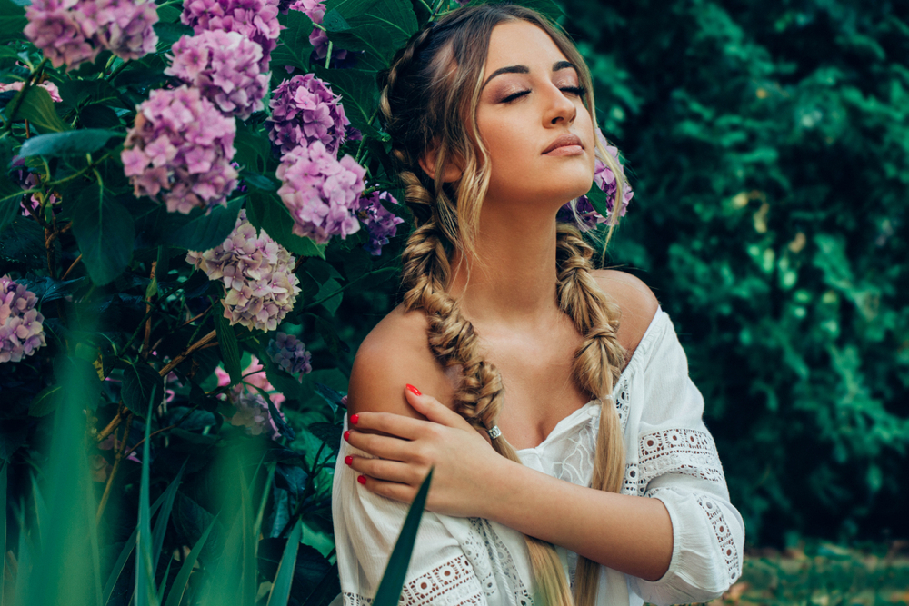 Girl closes her eyes and touches her shoulder outdoors with flowers behind her wearing a bubble braid hairstyle with fishtail braids