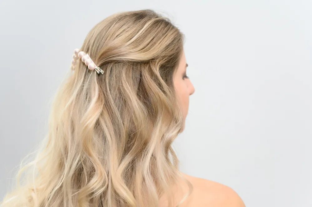 Blonde woman wears 2 pearl hair clips with wavy hair to model half up half down hairstyles in front of gray wall