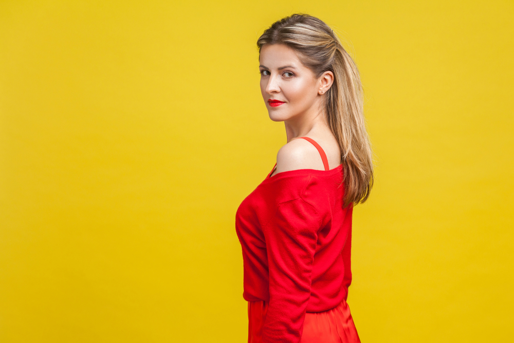 Woman with blonde highlights and brown hair looks over her shoulder with half up hairstyle in red dress with yellow background