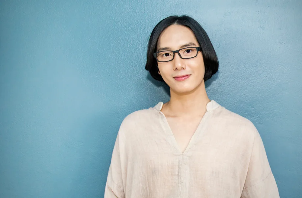Korean man stands in front of blue wall with collared cream colored shirt and long straight hairstyle