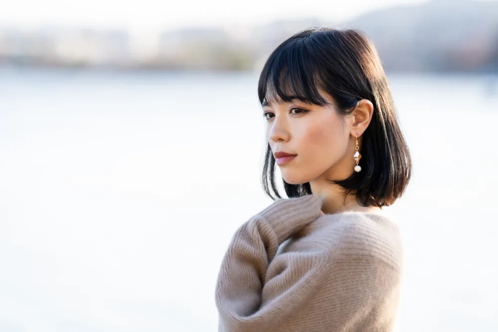 Asian woman in a long fuzzy sweater wears a Shoulder Length Lob With Wispy Blunt Bangs and holds her hand to her chest