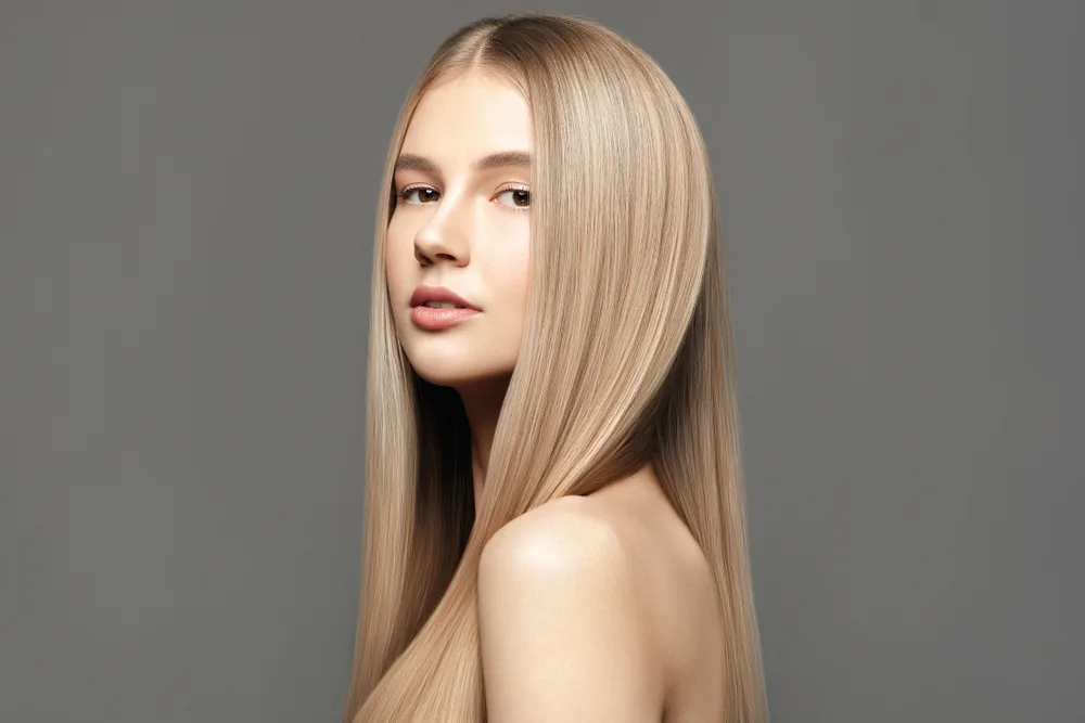Woman with bare shoulders in front of gray wall wears her hair straight in one of the best shades of blonde for neutral skin tones, beige blonde