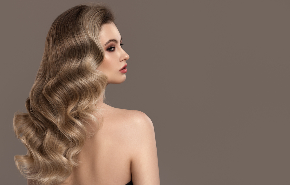 Back view of woman with bare shoulders modeling a mix between brown and shades of blonde with bronde hair color styled in waves