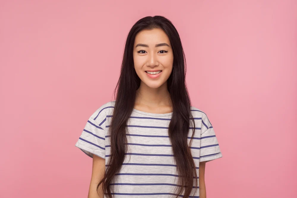 Young Asian long hair model with striped shirt smiling in front of pink wall