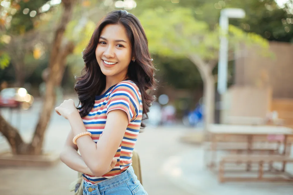 Young Asian woman walking through the park wears long hairstyle with waves and red and blue striped shirt
