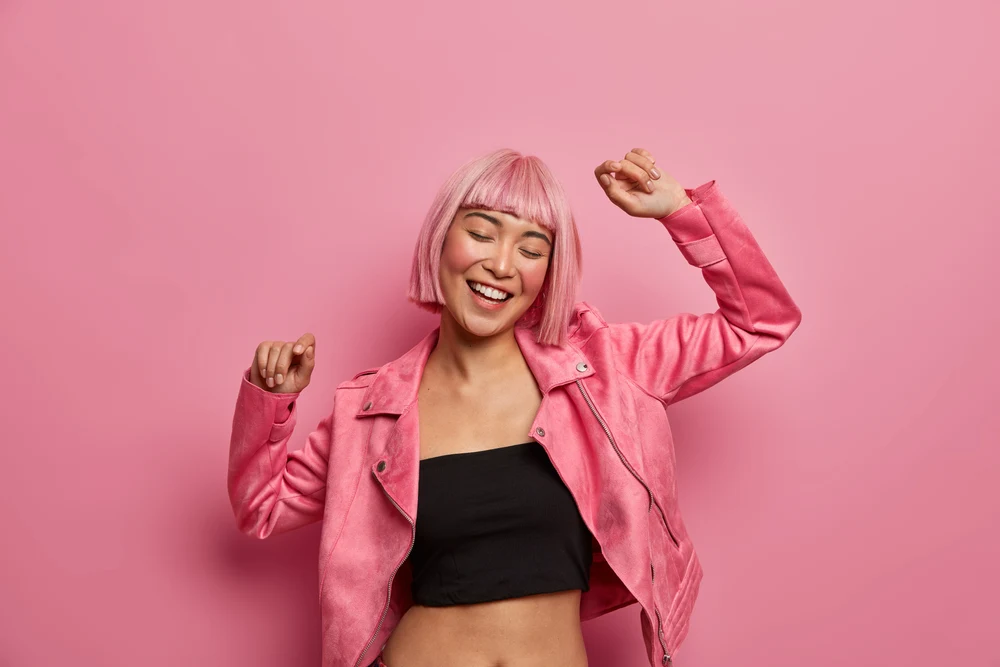 Young woman with light pink hair in a crop-top that is black in color wearing a pink leather jacket and holding her arms up dancing