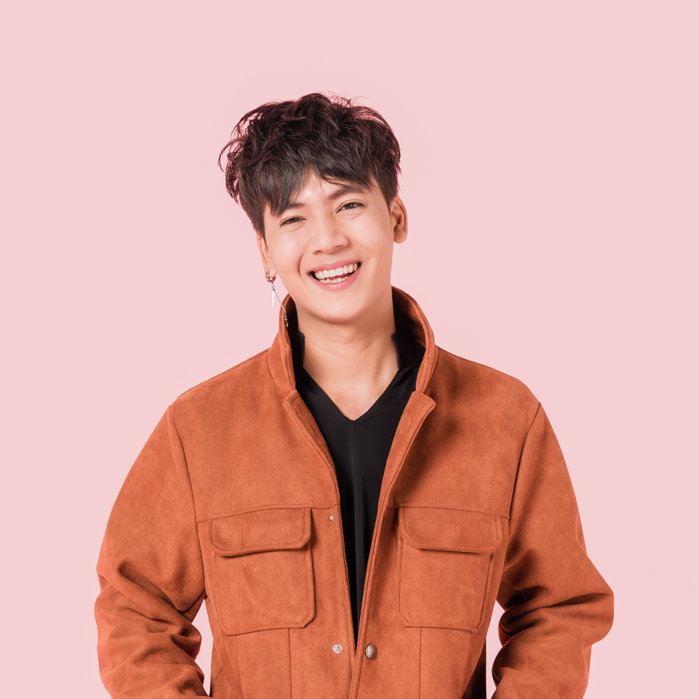 Young Korean man wears an orange jacket over a black shirt in front of pink wall to show off a messy volumized Korean hairstyle