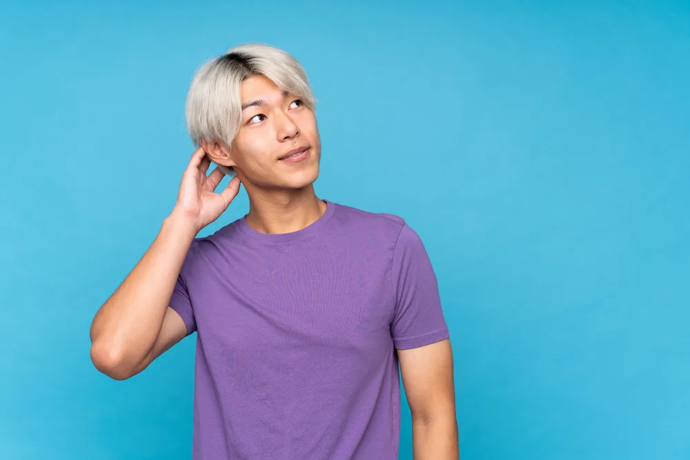 Asian man with platinum two block hairstyle in purple t-shirt stands in front of blue wall with hand on his head