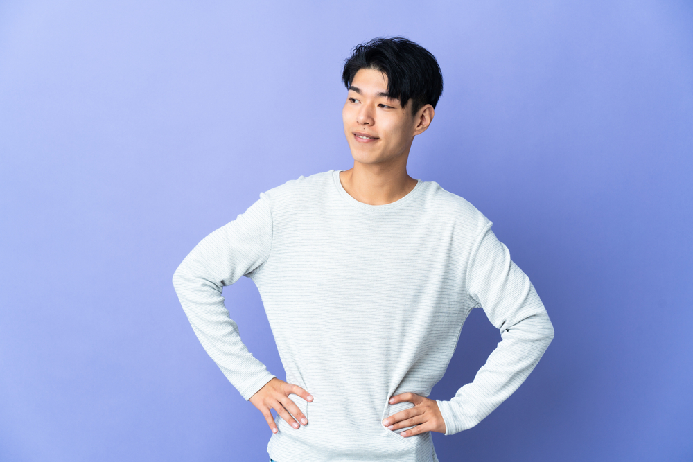 Young Asian man with hands on his hips poses in a white long sleeved shirt in front of a purple wall with short two block curtains hairstyle
