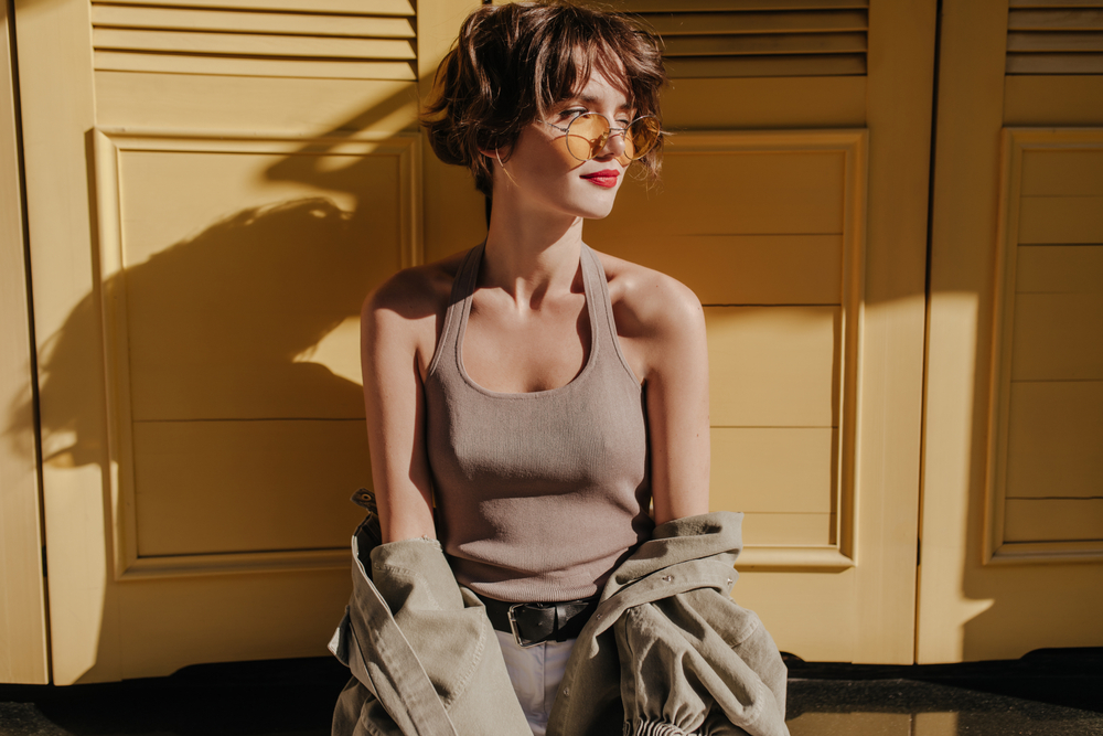 Woman sits in front of yellow doors with tank top and yellow sunglasses to show one of her favorite short heart shaped face hairstyles