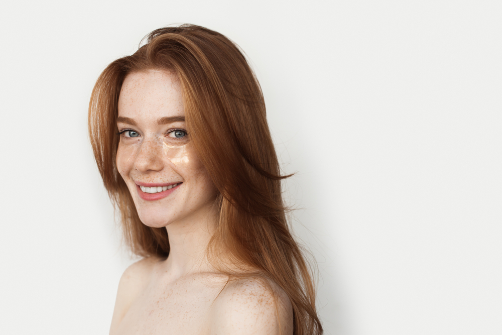 Red haired woman with bare shoulders wears one of the best heart shaped face haircuts with soft face-framing layers in long hair
