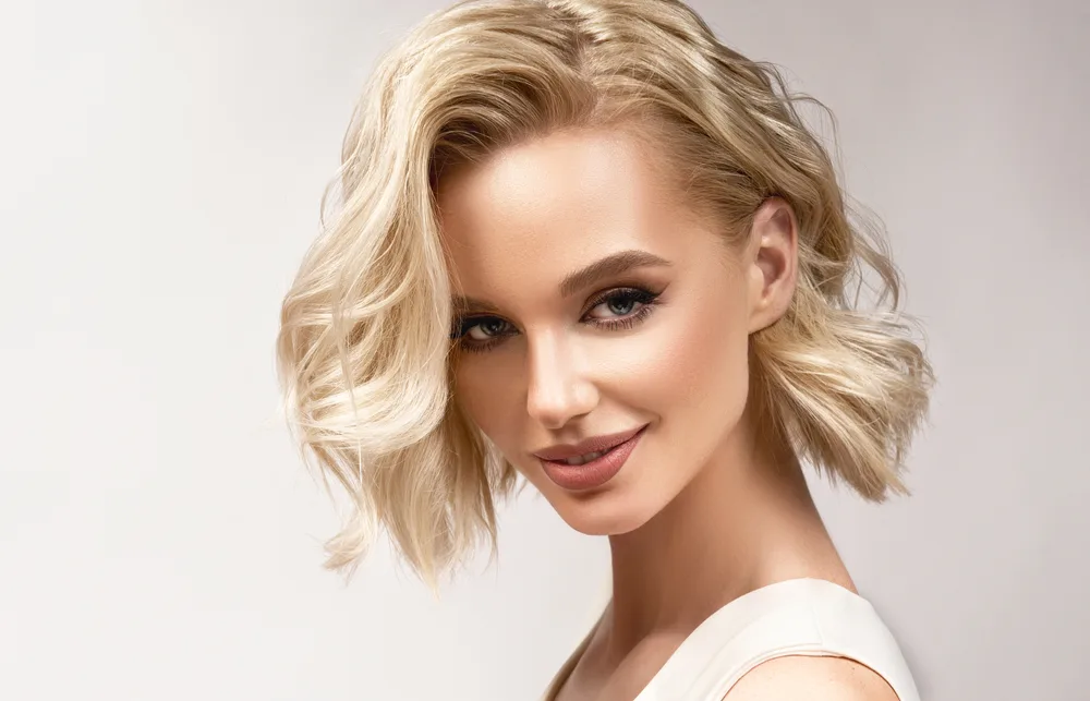 Blonde woman tilts her head and smiles as she wears one of many heart shaped face haircuts in a bob with side part and waves