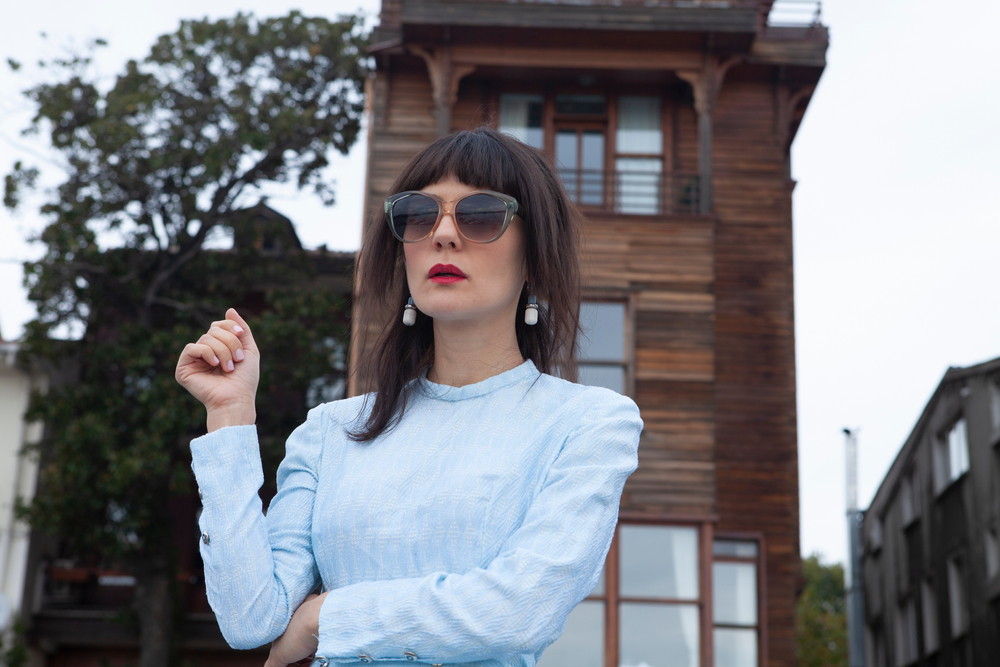 Brunette woman wears big sunglasses in front of wooden building with blue blouse to show examples of long shag haircuts