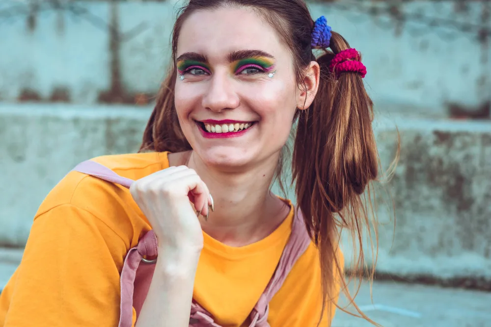 Young woman with hair styled in crazy pigtail buns wears bright eye makeup outside with orange shirt on