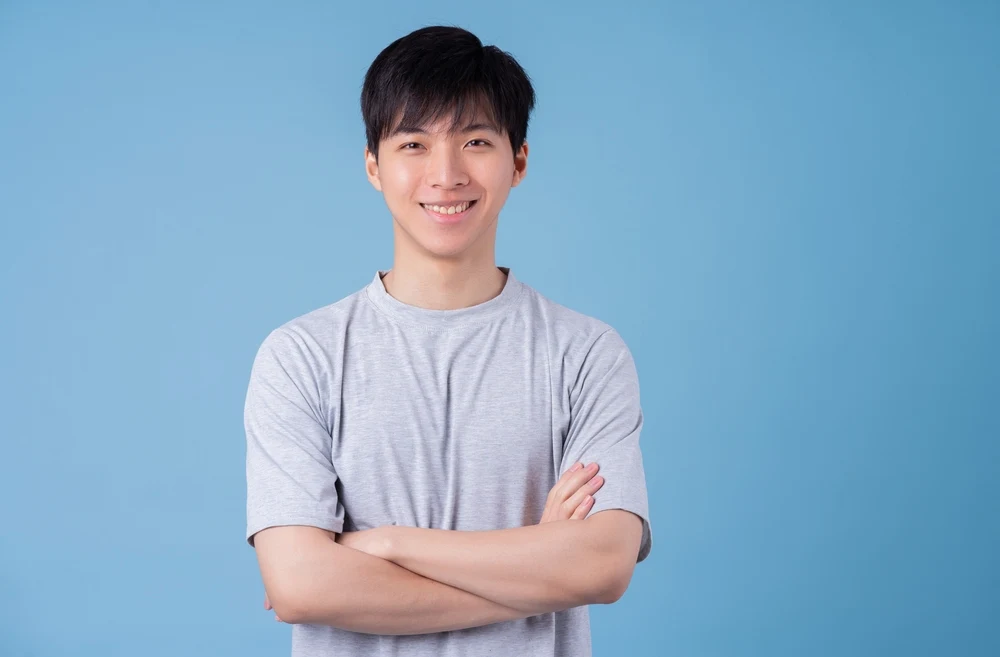 Man crosses his arms wearing a gray t-shirt in front of blue wall with straight hair brushed down over his forehead