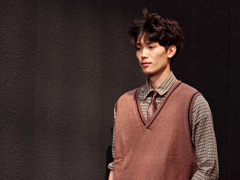 Korean man stands with hands in his pockets wearing a sweater vest and collared shirt with a mushroom cut styled in a blowout