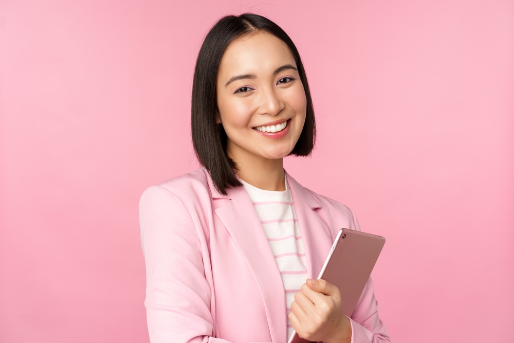 Smiling woman with dark hair holds a tablet in pink blazer in front of pink wall to show her short haircut on a heart shaped face