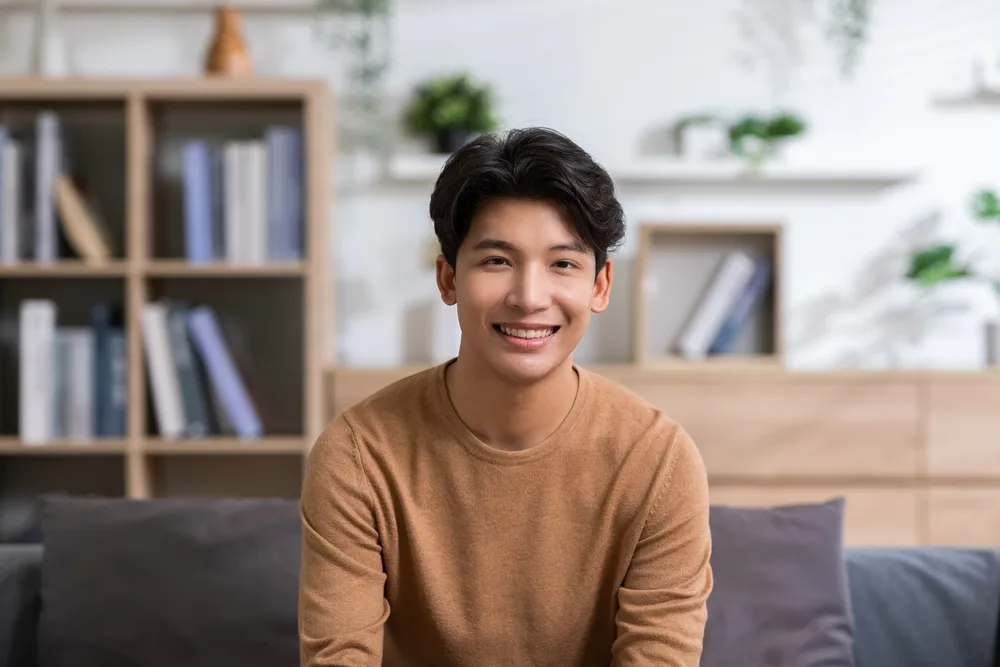 Young Korean man smiles and sits on couch with tan sweater and long, softly textured hairstyle