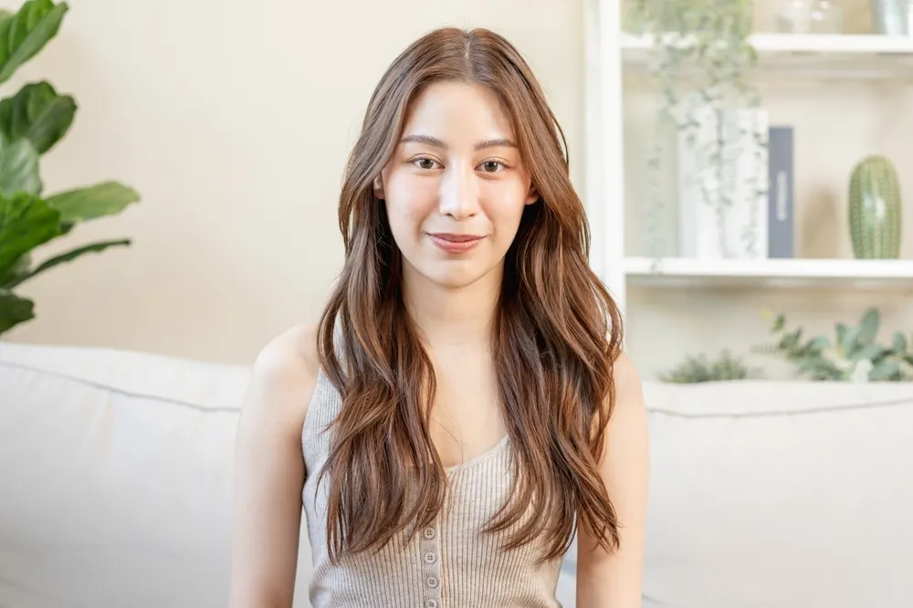 Smiling woman with Asian long hair style wears waves and a middle part while sitting on the couch near a bookshelf