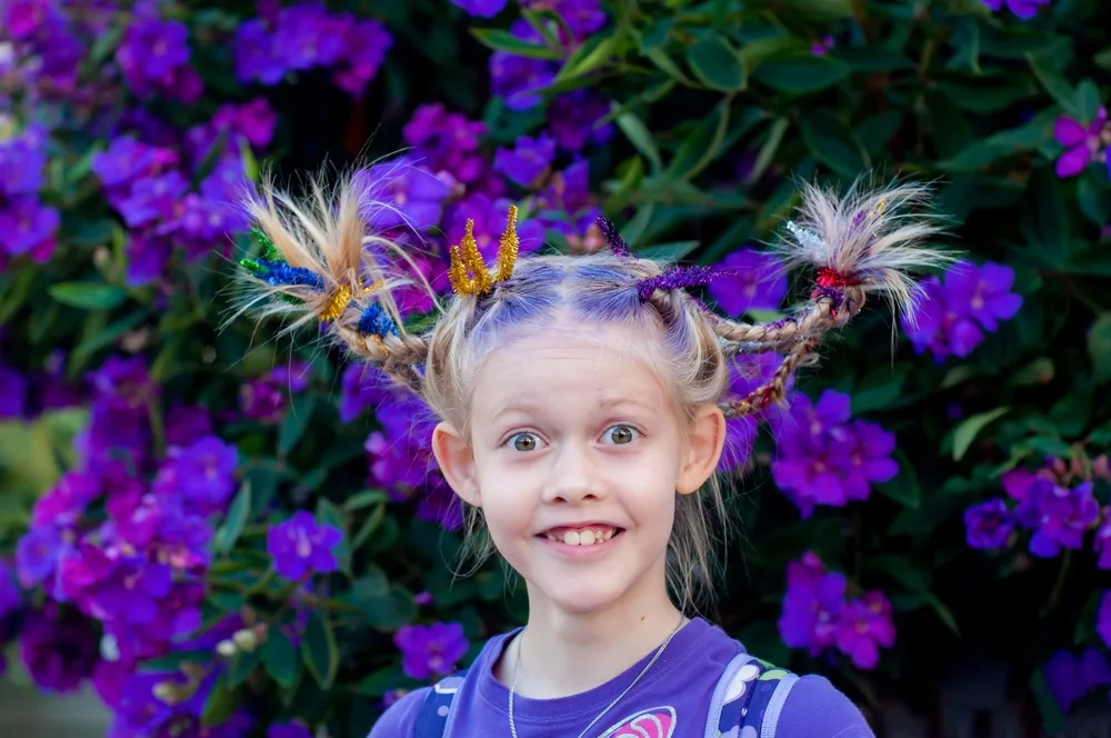 Little girl wears pigtail braids with colorful pipe cleaners shaping them in front of purple flower bushes 