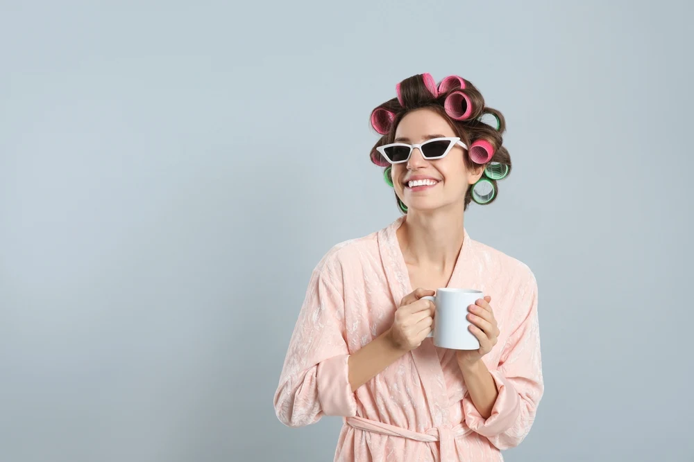 Woman in bathrobe wears curlers in her hair for crazy hair day ideas with white sunglasses and a mug while smiling in front of gray wall