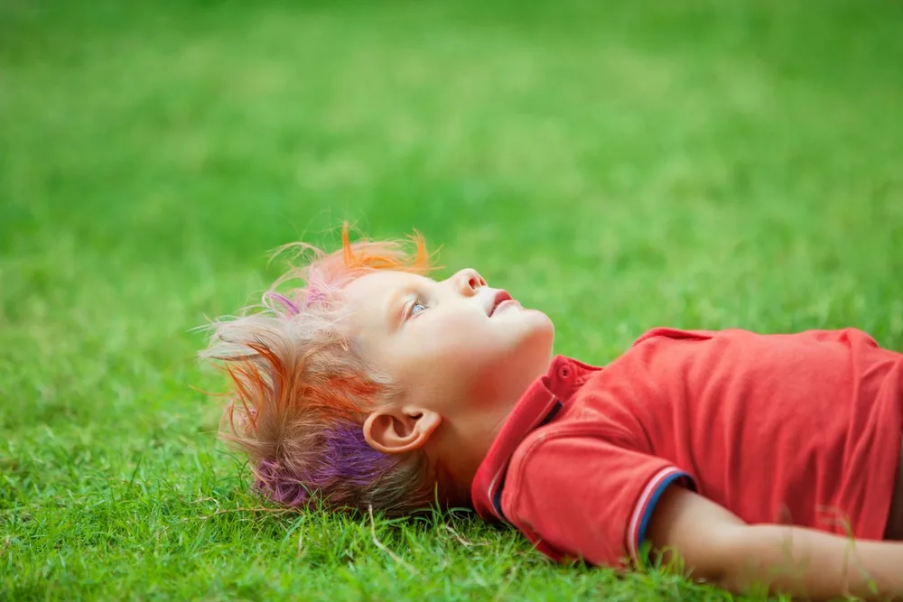 Little boy with orange and purple colored hair wears one of the crazy hair day ideas with messy style while laying on the grass