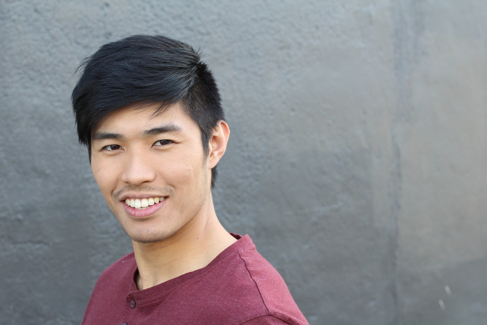 Asian man smiles wearing a red shirt in front of gray wall to show off his hairstyle with straight, layered hair styled to the side