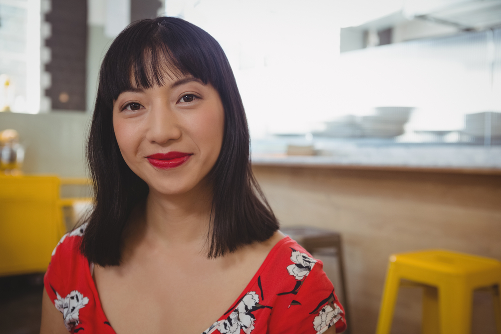 Blunt Shoulder Length Cut With Piece-y Bangs on an Asian woman in a red floral shirt sitting in front of a window