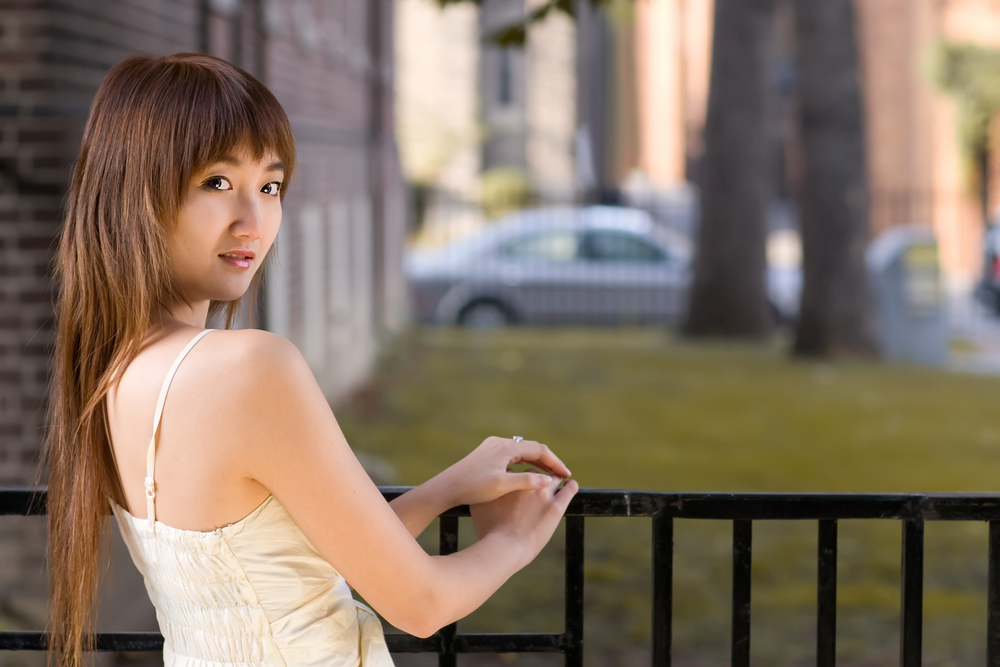Young Asian woman stands at railing outside looking over her shoulder with white dress showing her version of long shag haircuts